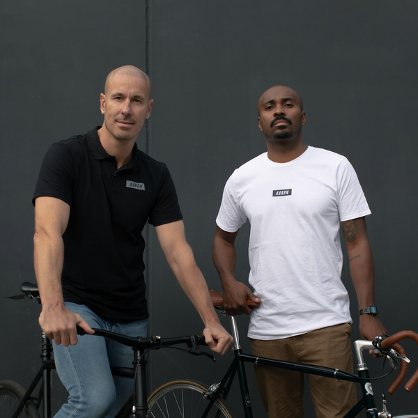 Zwei Männer stehen mit Fahrrädern vor einer dunkelgrauen Wand. Der eine trägt ein schwarzes Polohemd und Jeans und sitzt auf dem Fahrrad, der andere trägt ein weißes T-Shirt und eine beigefarbene Hose und steht neben seinem Fahrrad. Beide blicken selbstbewusst in die Kamera.