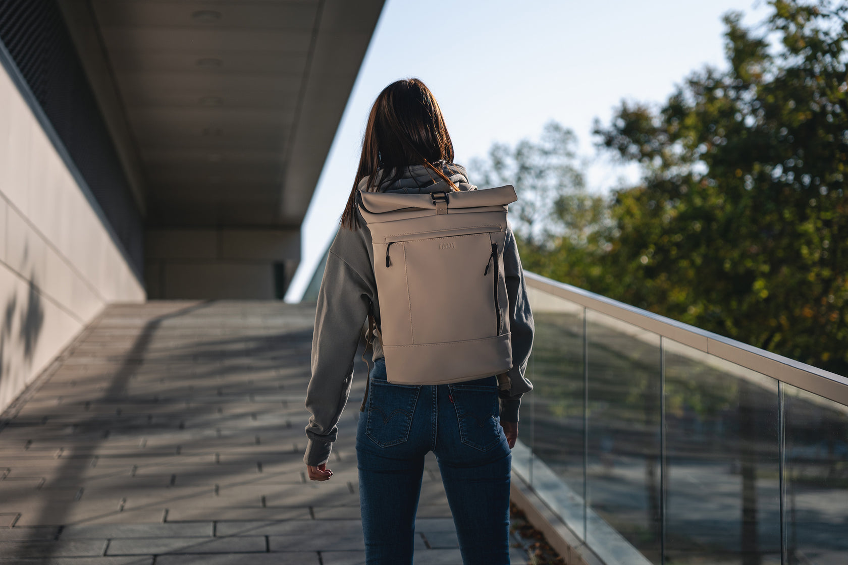 Eine Person mit langen Haaren, die einen grauen Kapuzenpullover und Jeans trägt, geht draußen auf einem gepflasterten Weg neben einem Glasgeländer und trägt einen hellen Rucksack. Im Hintergrund sind Bäume und ein modernes Gebäude zu sehen.