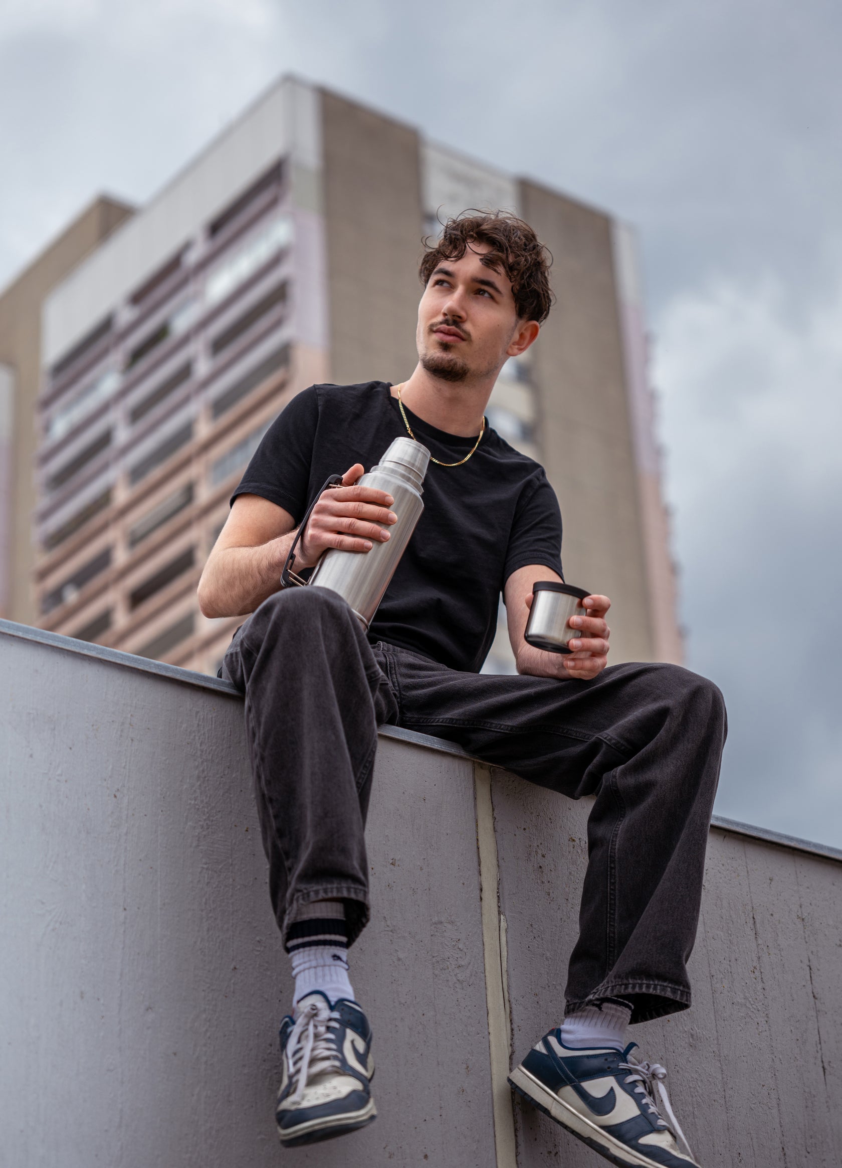 Ein junger Mann in schwarzem T-Shirt und Jeans sitzt auf einem Dachvorsprung mit einer Metallthermoskanne und einer Tasse in der Hand, mit hohen Gebäuden und einem wolkenverhangenen Himmel im Hintergrund.