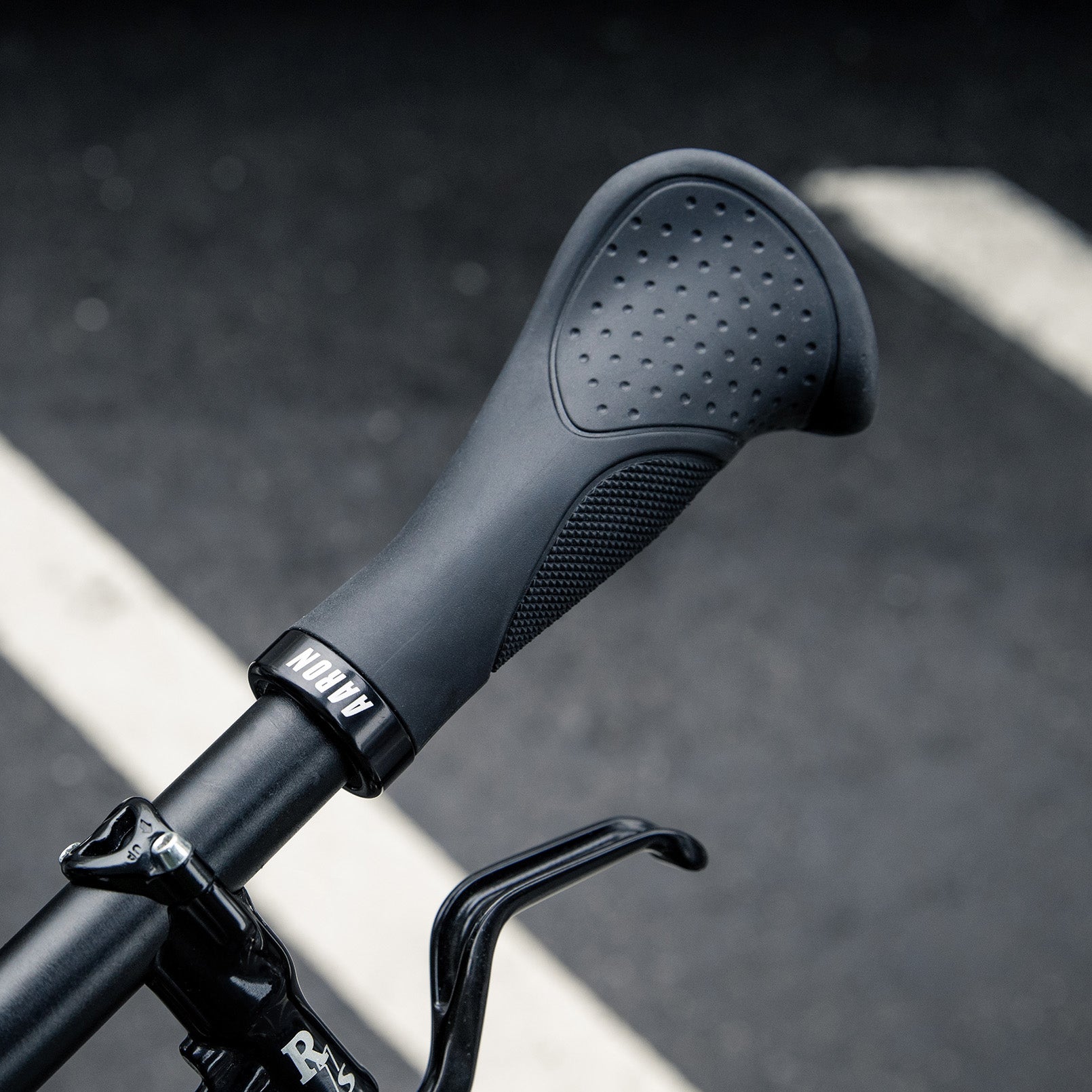 Nahaufnahme eines schwarzen ergonomischen Fahrradlenkergriffs mit strukturierter Oberfläche und ARION-Schriftzug, der an einem Fahrrad befestigt ist, vor einem unscharfen Straßenhintergrund, auf dem weiße Straßenlinien zu sehen sind.