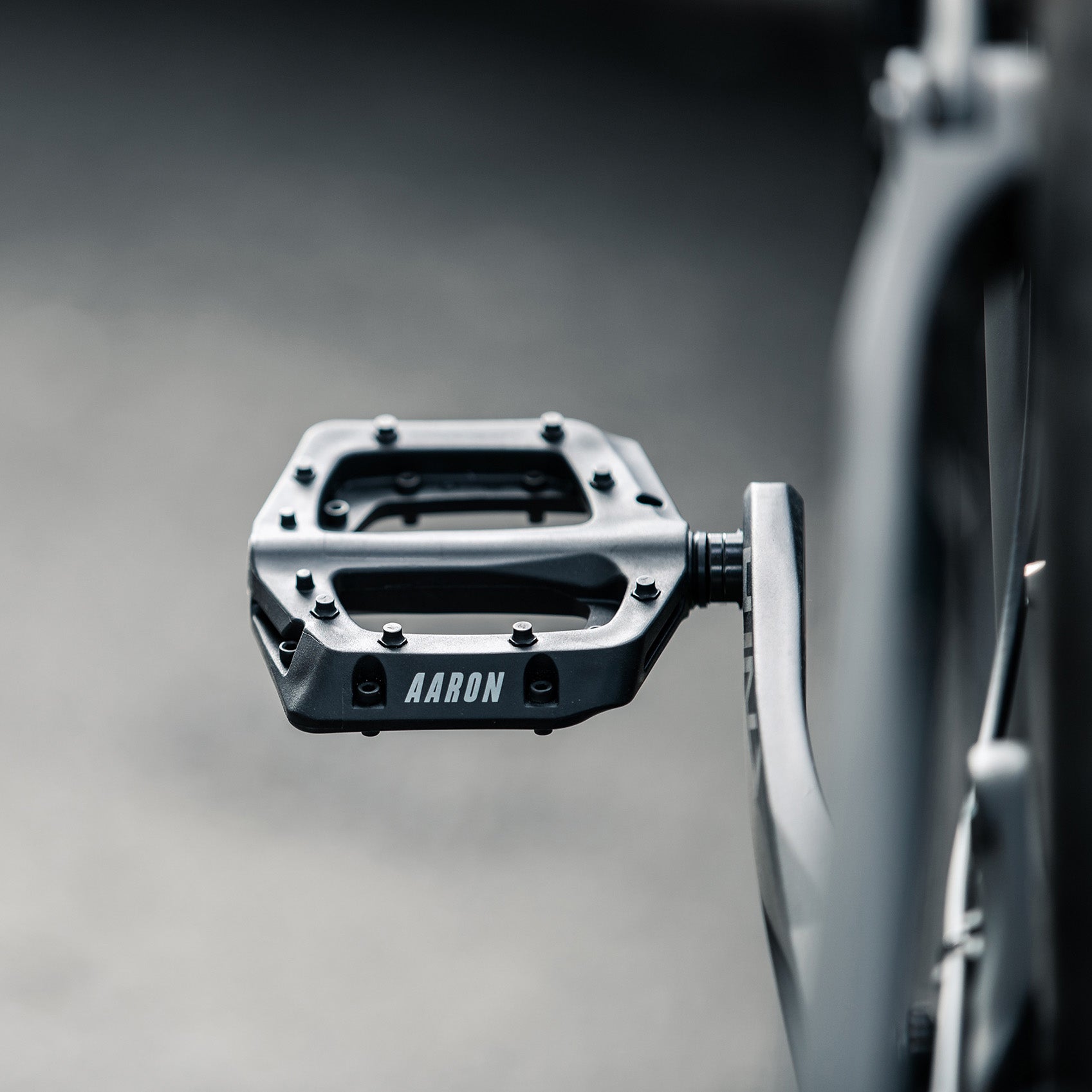 Nahaufnahme eines schwarzen Fahrradpedals aus Metall mit dem aufgedruckten Wort AARON, das an der Kurbel des Fahrrads befestigt ist, vor einem unscharfen, neutralen Hintergrund.