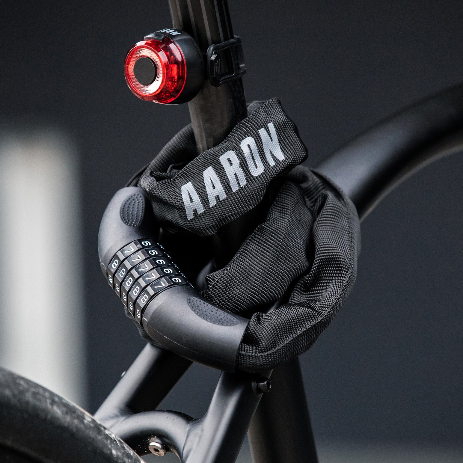 Ein schwarzes Fahrradschloss mit einer Stoffabdeckung mit der Aufschrift AARON sichert einen Fahrradrahmen. Ein rotes Fahrradrücklicht ist an der Sattelstütze befestigt, und der Hintergrund ist unscharf.