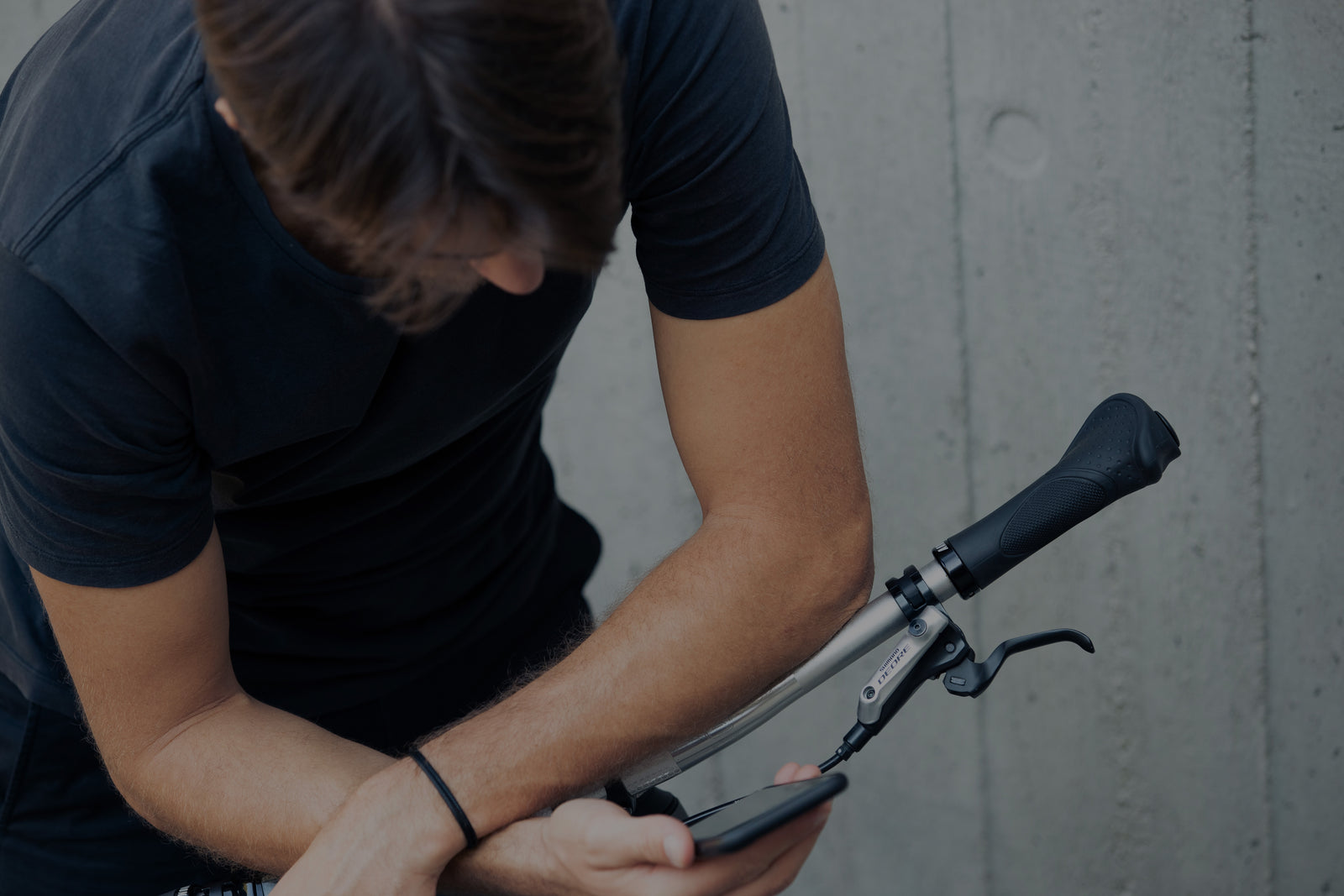 Eine Person in einem schwarzen T-Shirt blickt auf ein Smartphone, während sie den Lenker eines Fahrrads hält; der Hintergrund ist eine graue Betonwand.