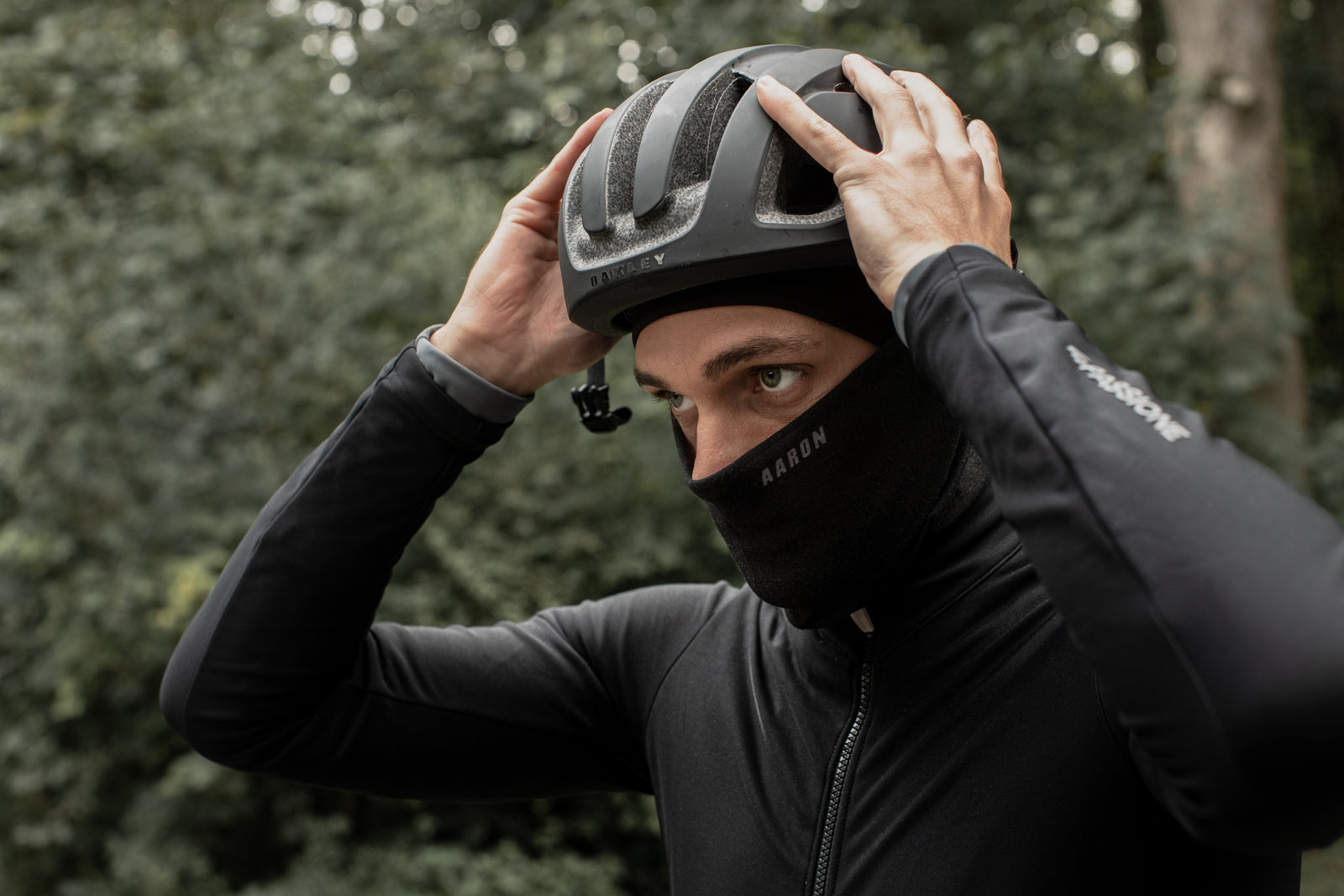 Eine Person, die eine schwarze Jacke und eine Sturmhaube trägt, passt einen schwarzen Fahrradhelm im Freien an, mit grünen Bäumen im Hintergrund.