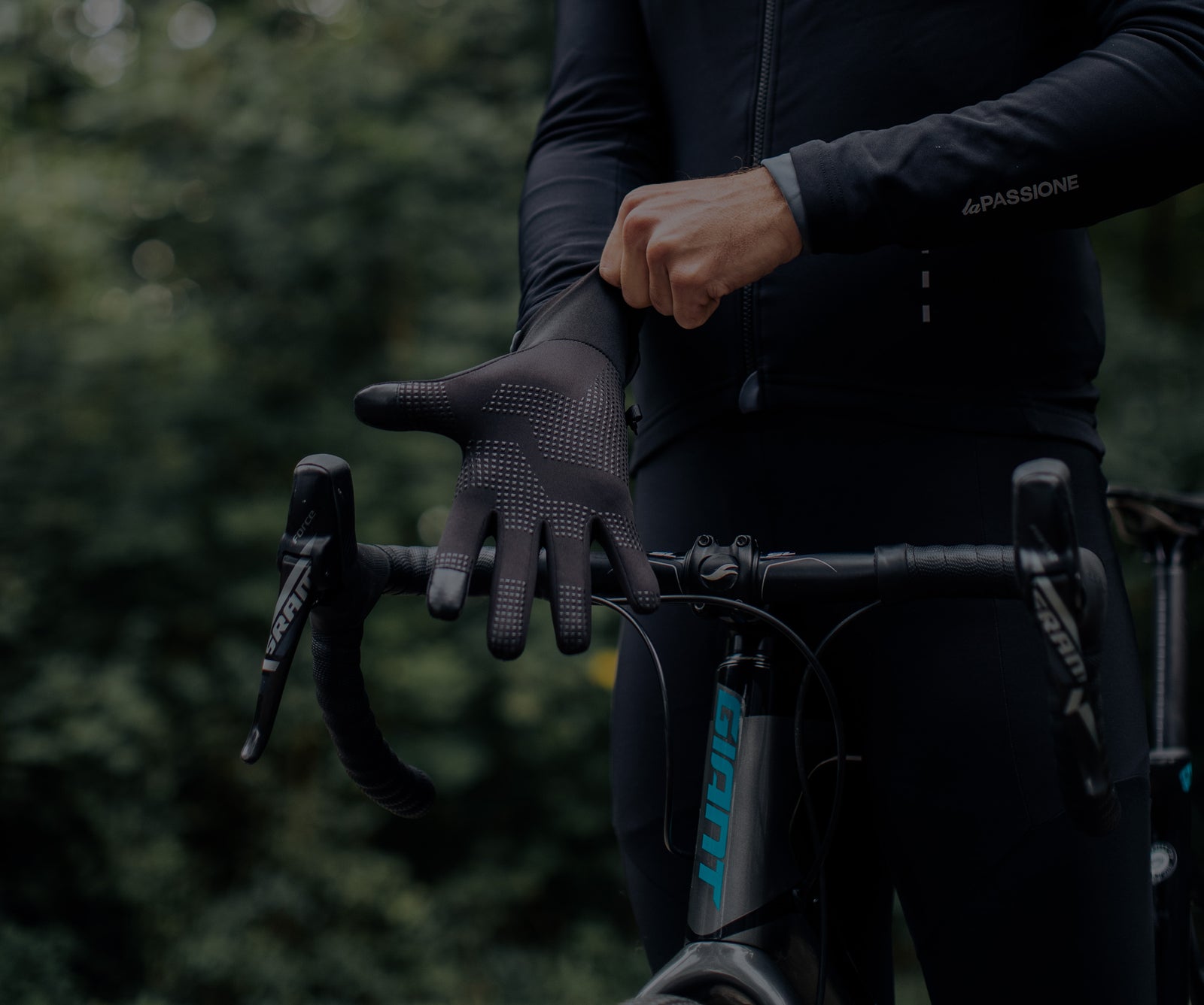 Ein Radfahrer in dunkler Kleidung steht im Freien und zieht einen schwarzen Handschuh an, während er den Lenker eines Fahrrads festhält, mit grünem Laub im verschwommenen Hintergrund.