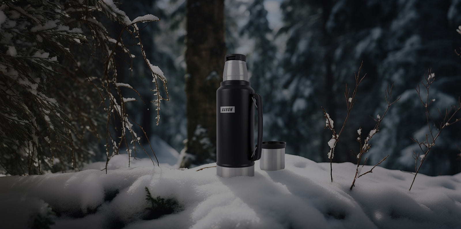 Eine schwarze Thermoskanne mit einem silbernen Becher steht auf dem schneebedeckten Boden in einem verschneiten Wald, umgeben von Bäumen und Ästen.
