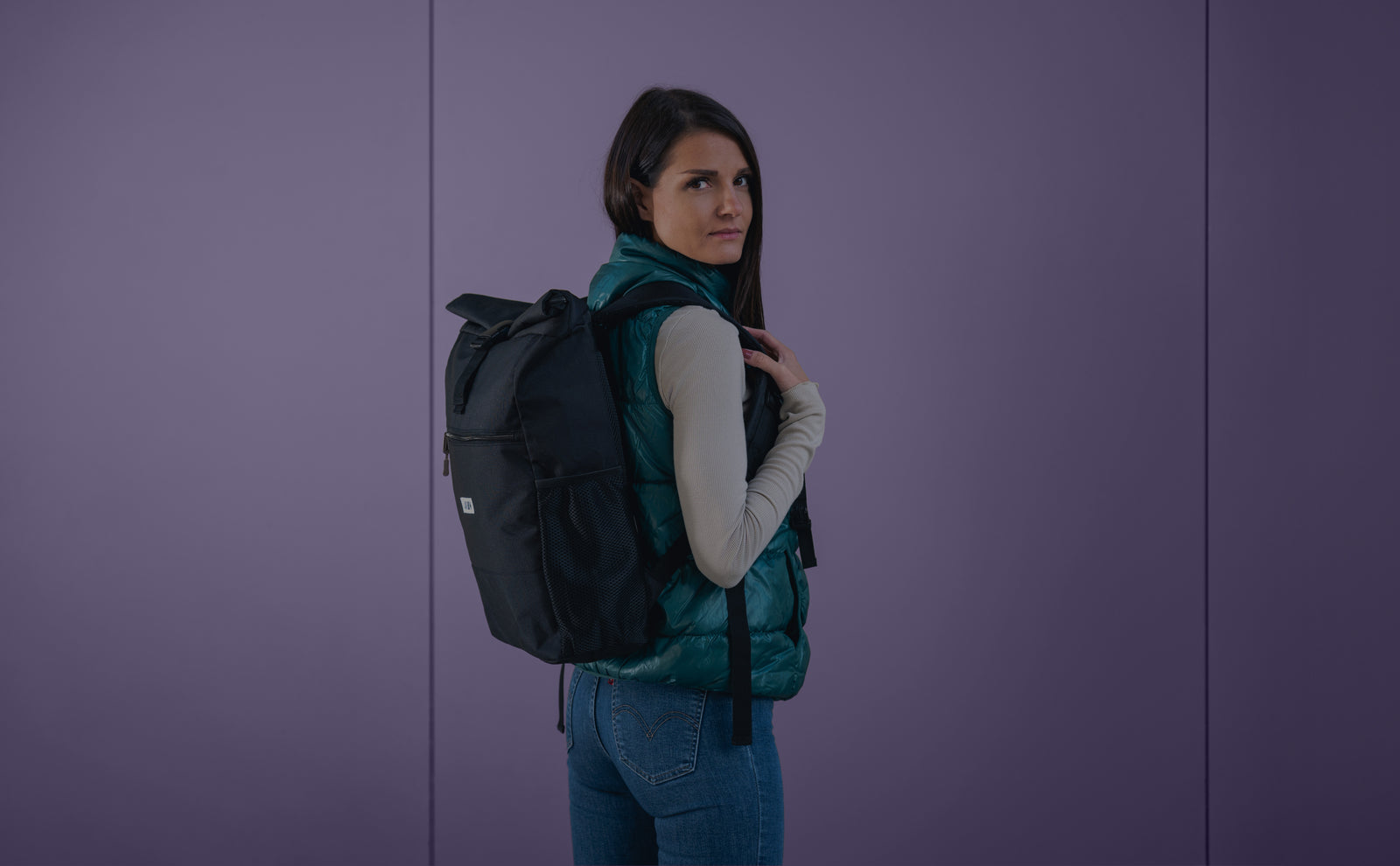 Eine Frau mit langen dunklen Haaren, die eine grüne Weste, ein beigefarbenes langärmeliges Hemd und blaue Jeans trägt, steht seitlich mit einem schwarzen Rucksack über der Schulter vor einem einfarbigen lila Hintergrund.